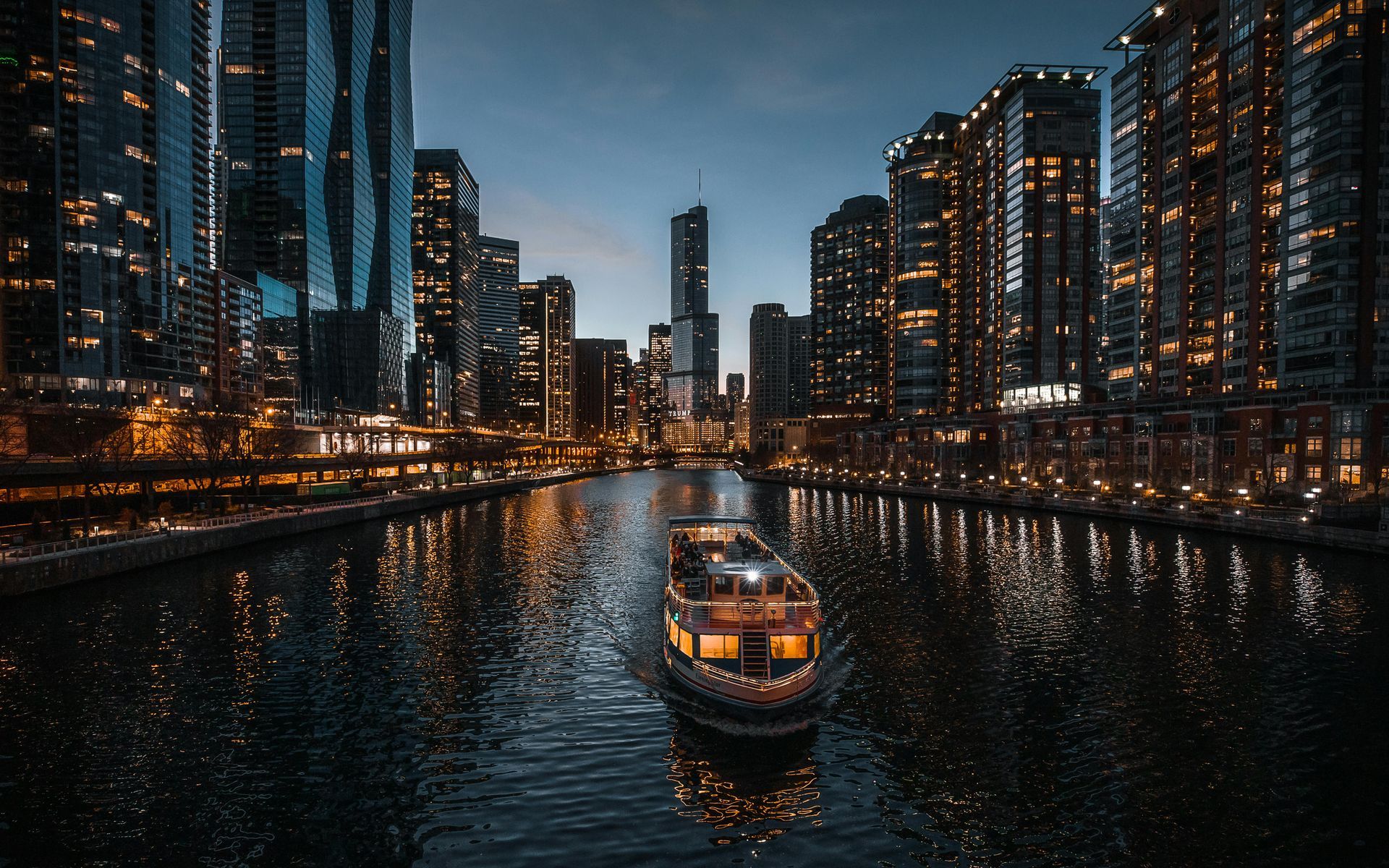 Boat on a dark river, flanked by illuminated skyscrapers at dusk.