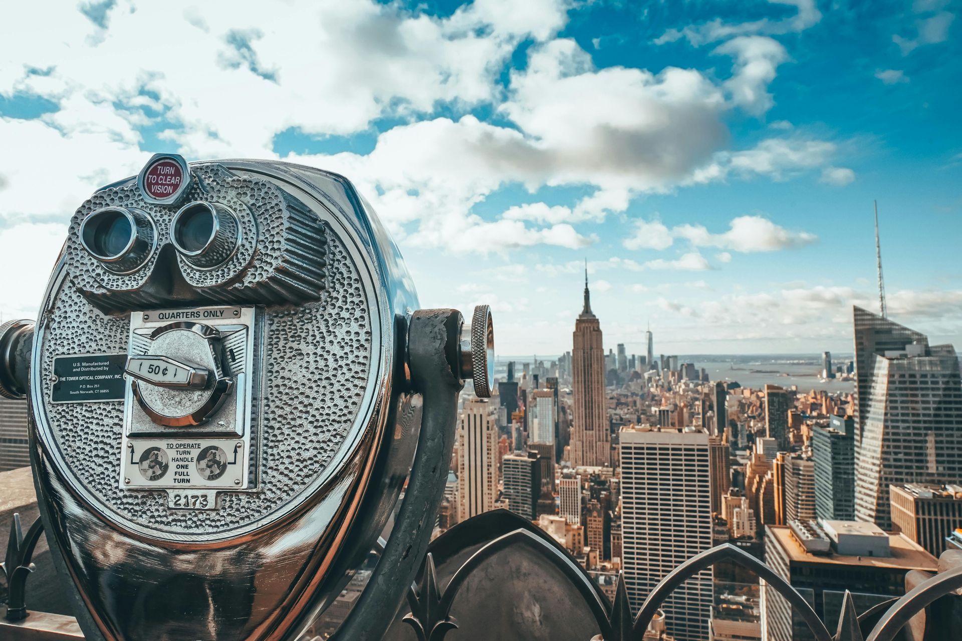 Binoculars overlooking the New York City skyline, including the Empire State Building, under a partly cloudy sky.