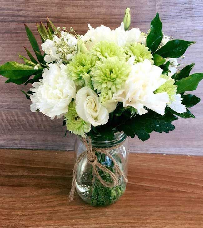 A Mason Jar Filled With White and Green Flowers is Sitting on a Wooden Table — Flower Hunt in Robina, QLD