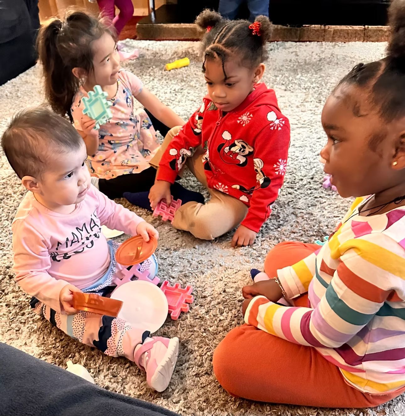 Four children sit on a gray rug, gathered closely and playing with toys indoors.