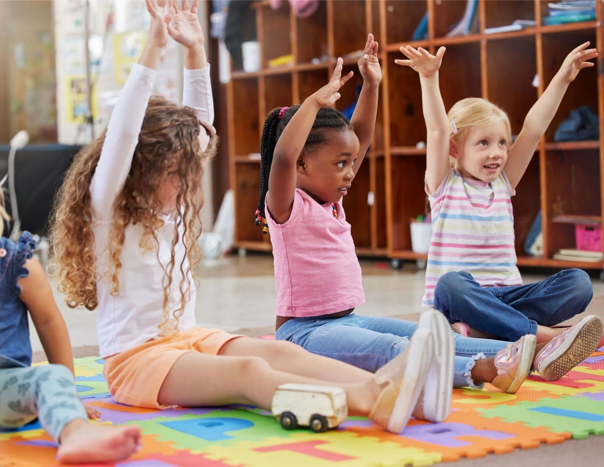 Children seated on colorful mats with arms raised during a classroom activity