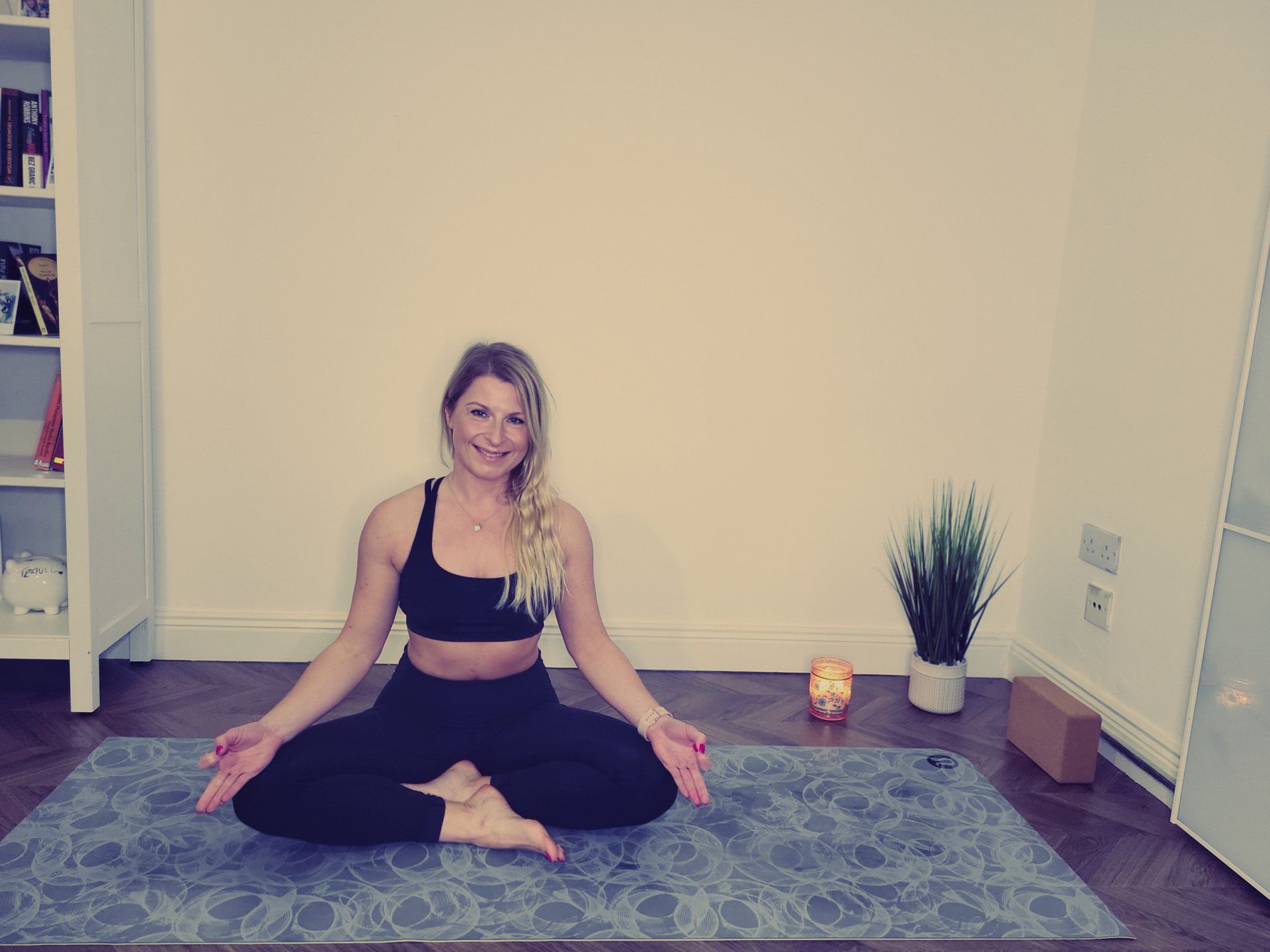 Woman seated in yoga pose on mat, smiling. Indoor setting with plant, block, and bookcase.