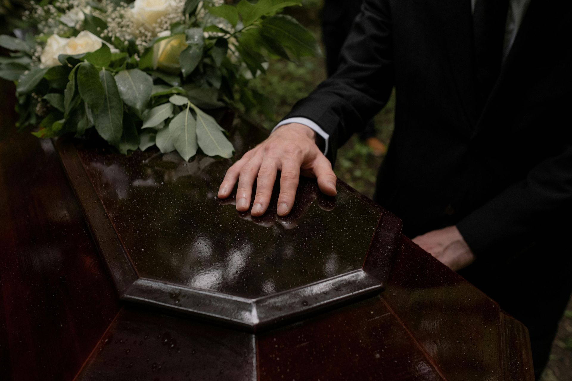 Casket with a hand resting on it beside white flowers at a funeral service