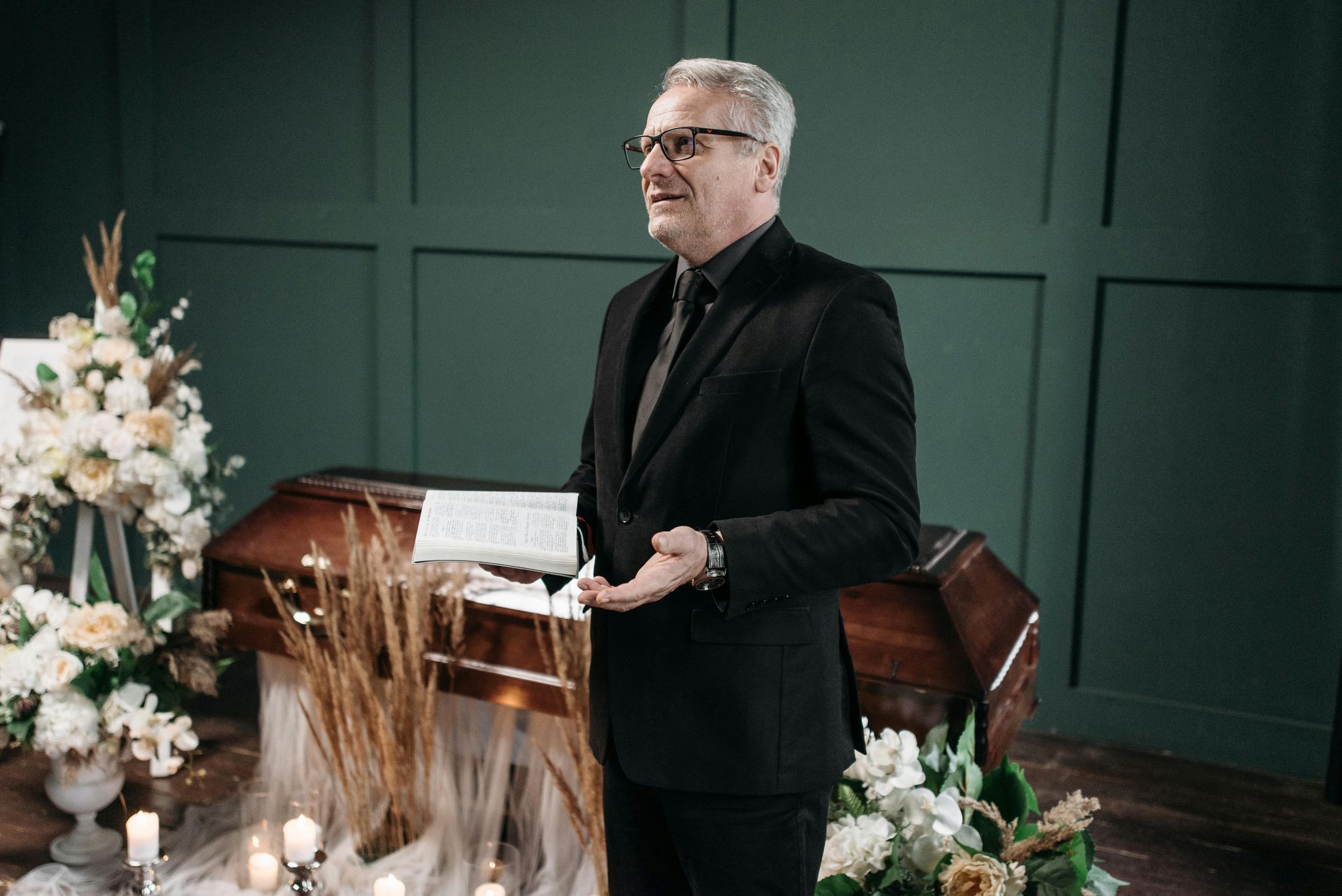 Man in a black suit speaking beside a casket at a funeral, with flowers and green paneled walls behind him