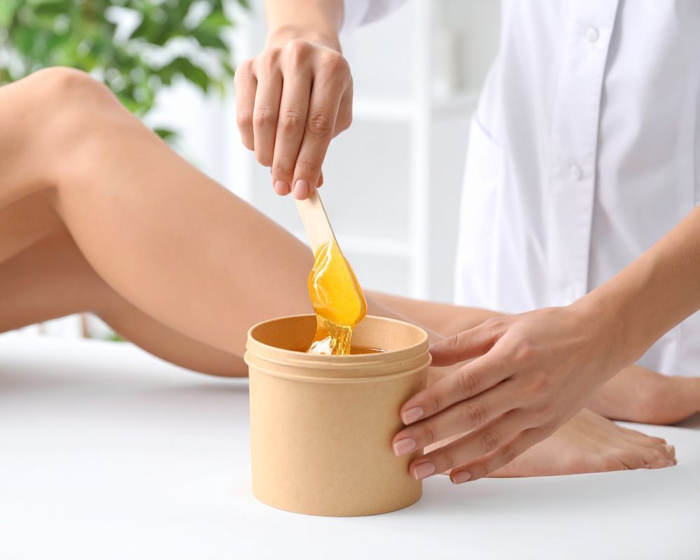 Person applying wax to a woman's leg with a wooden spatula. Jar of wax on a table.