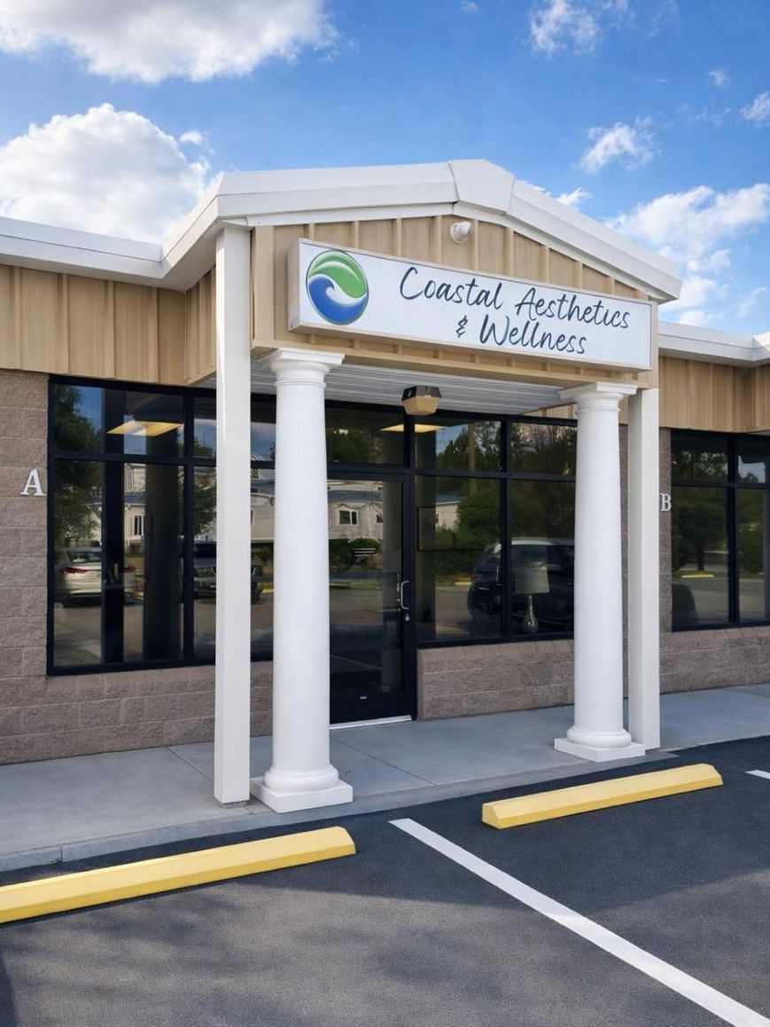 Entrance to Coastal Aesthetics & Wellness with white pillars, a glass front door, and tan siding under a blue sky.