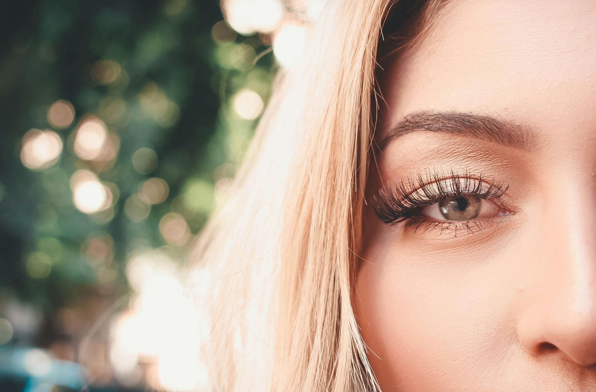 Close-up of a person's eye with glittery makeup and long lashes, set against a blurred, sunlit outdoor background.