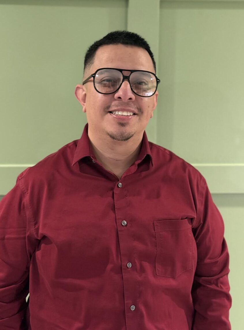 Man with glasses and a red shirt smiling in front of a green wall.