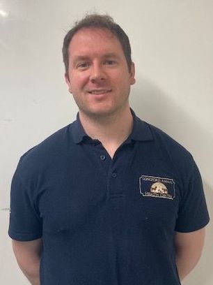 A man wearing a blue polo shirt is standing in front of a white wall.