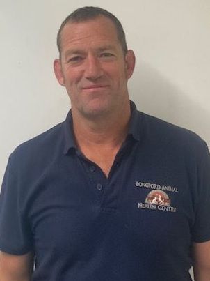 A man wearing a blue polo shirt is standing in front of a white wall.