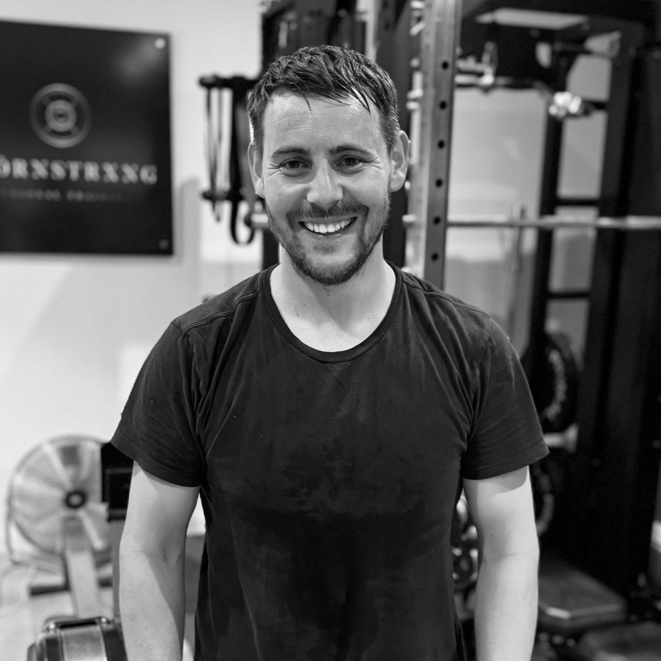 Man in black t-shirt smiles in gym, equipment visible in background.