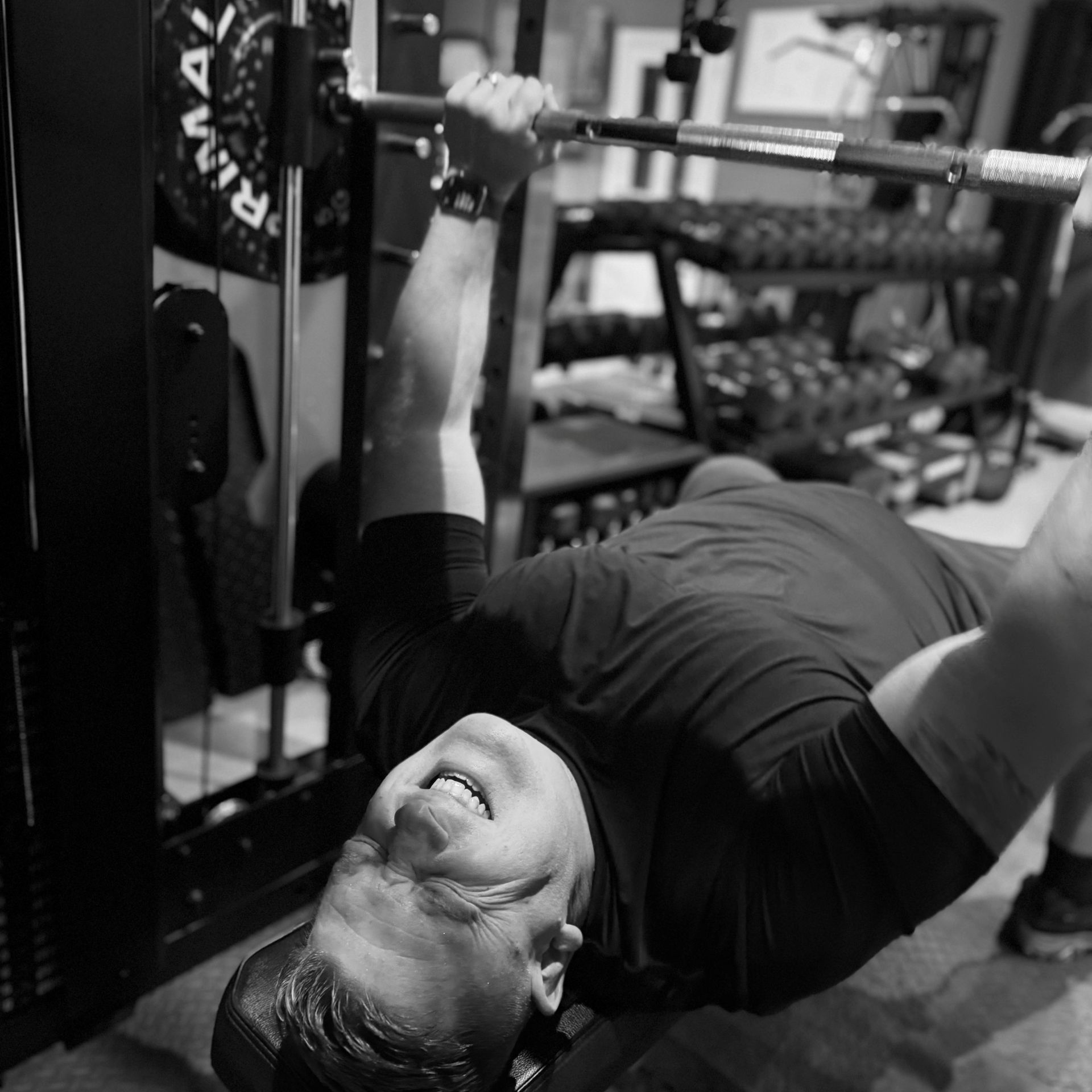 Man bench pressing in home gym, straining facial muscles, barbell, weights in background.