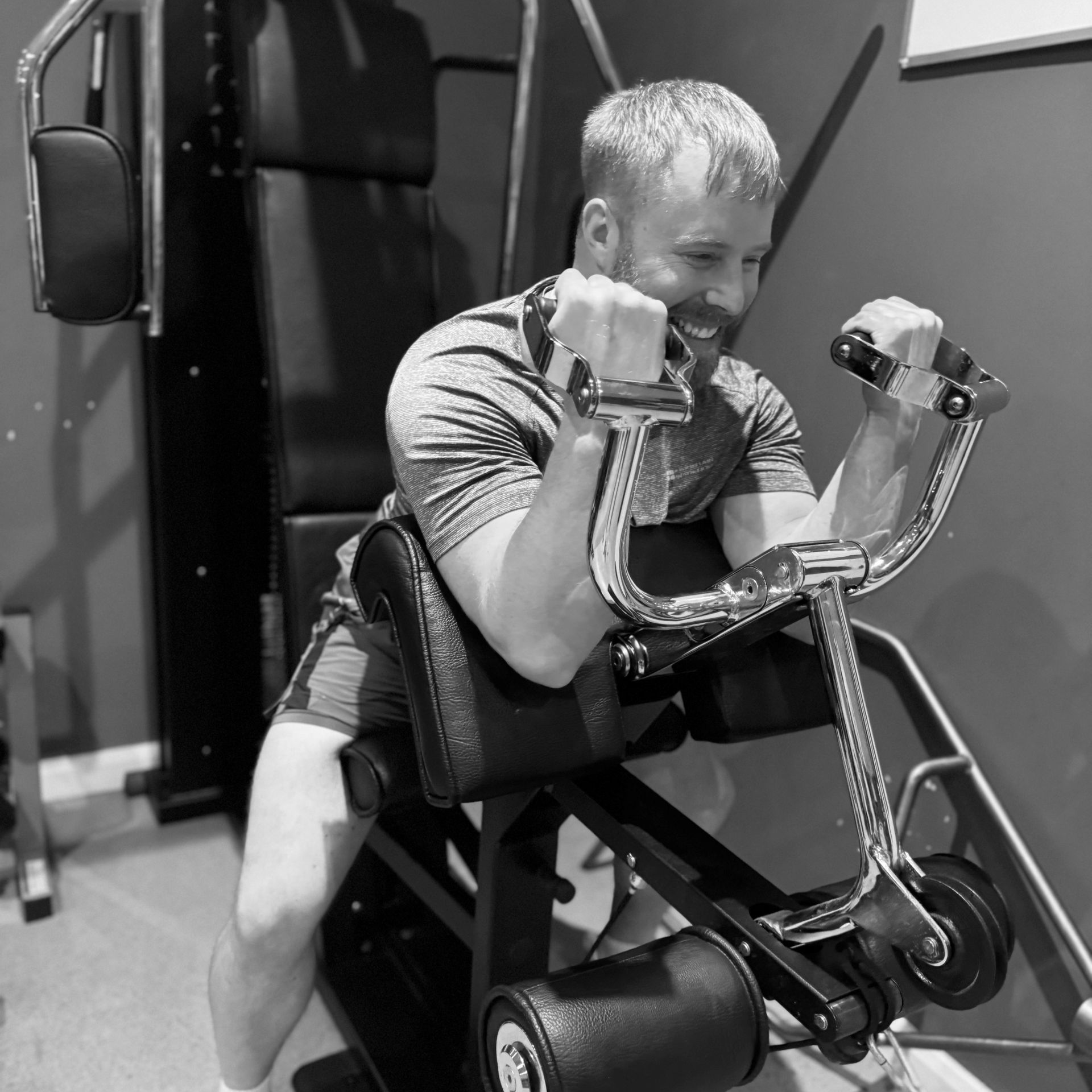 Man using a bicep curl machine, indoors. He's flexing with a focused expression. Black and white.
