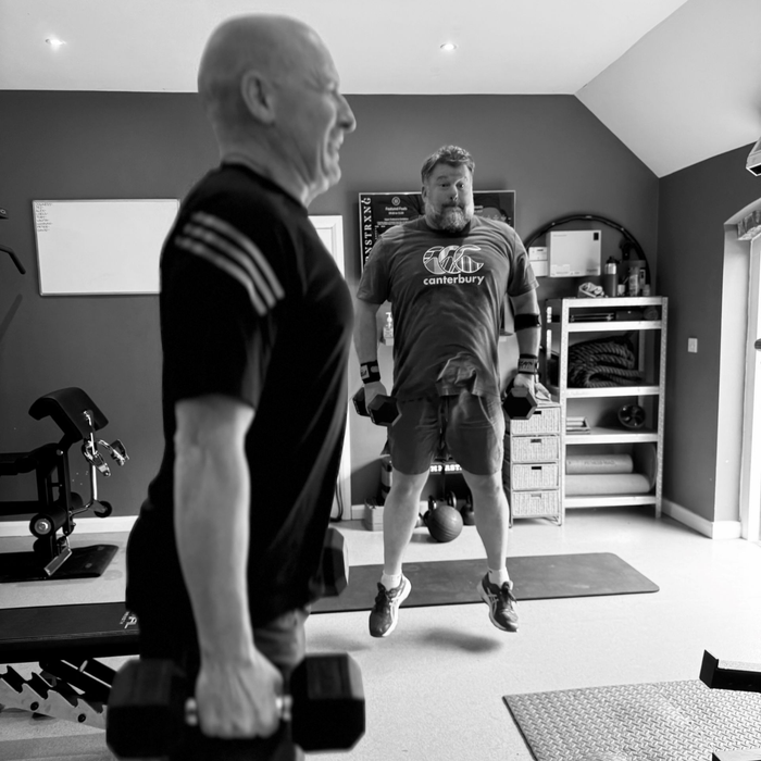 Two men exercising in a home gym, one lifting weights, the other jumping with dumbbells.