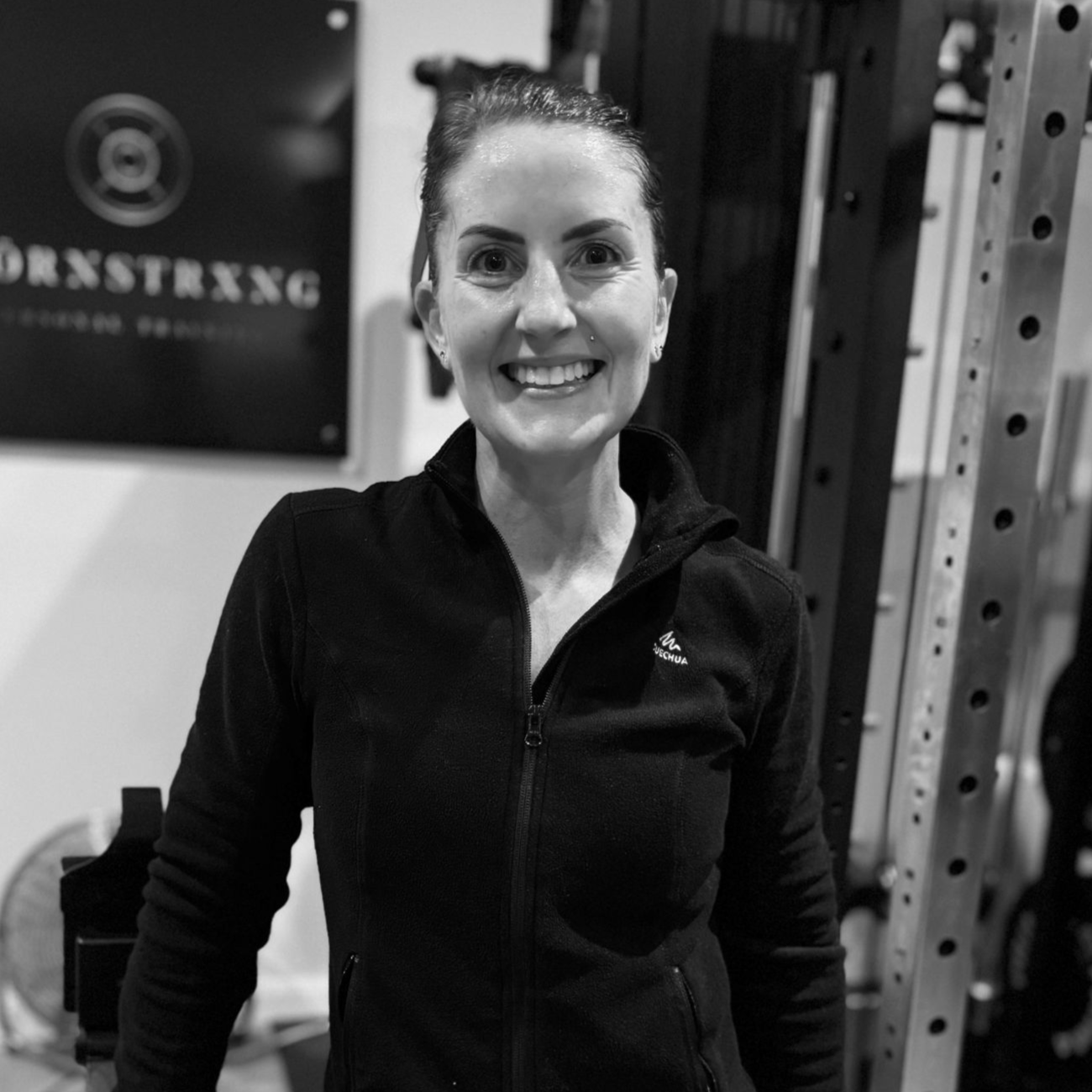Woman smiling, wearing a black zip-up jacket, standing in a gym.