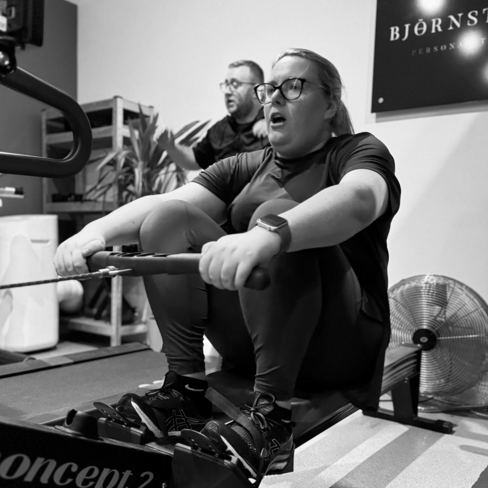 Woman rowing indoors, grimacing, Concept2 machine. Man in background. Black and white.