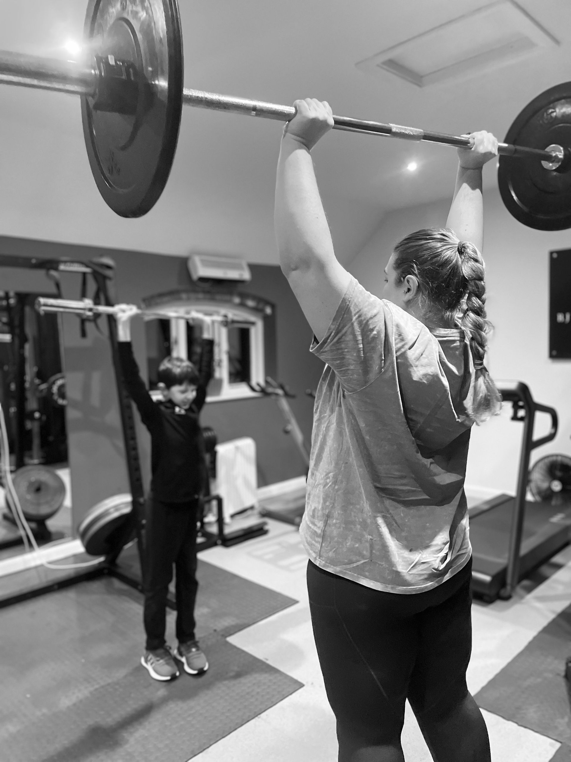 Woman and child lift barbells overhead in a home gym, focus on their backs.