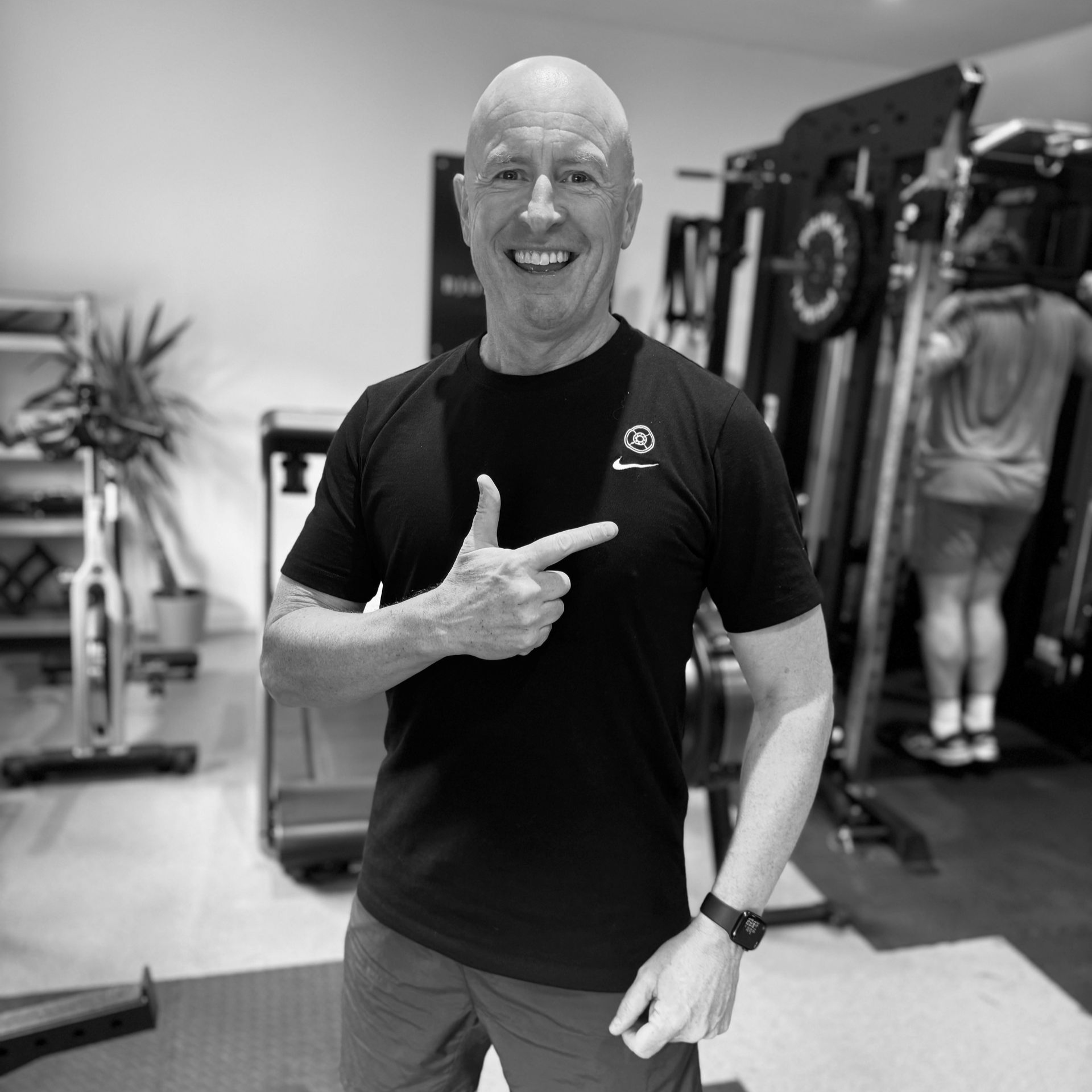 Man in gym points to the right; wearing black shirt, watch, and shorts.