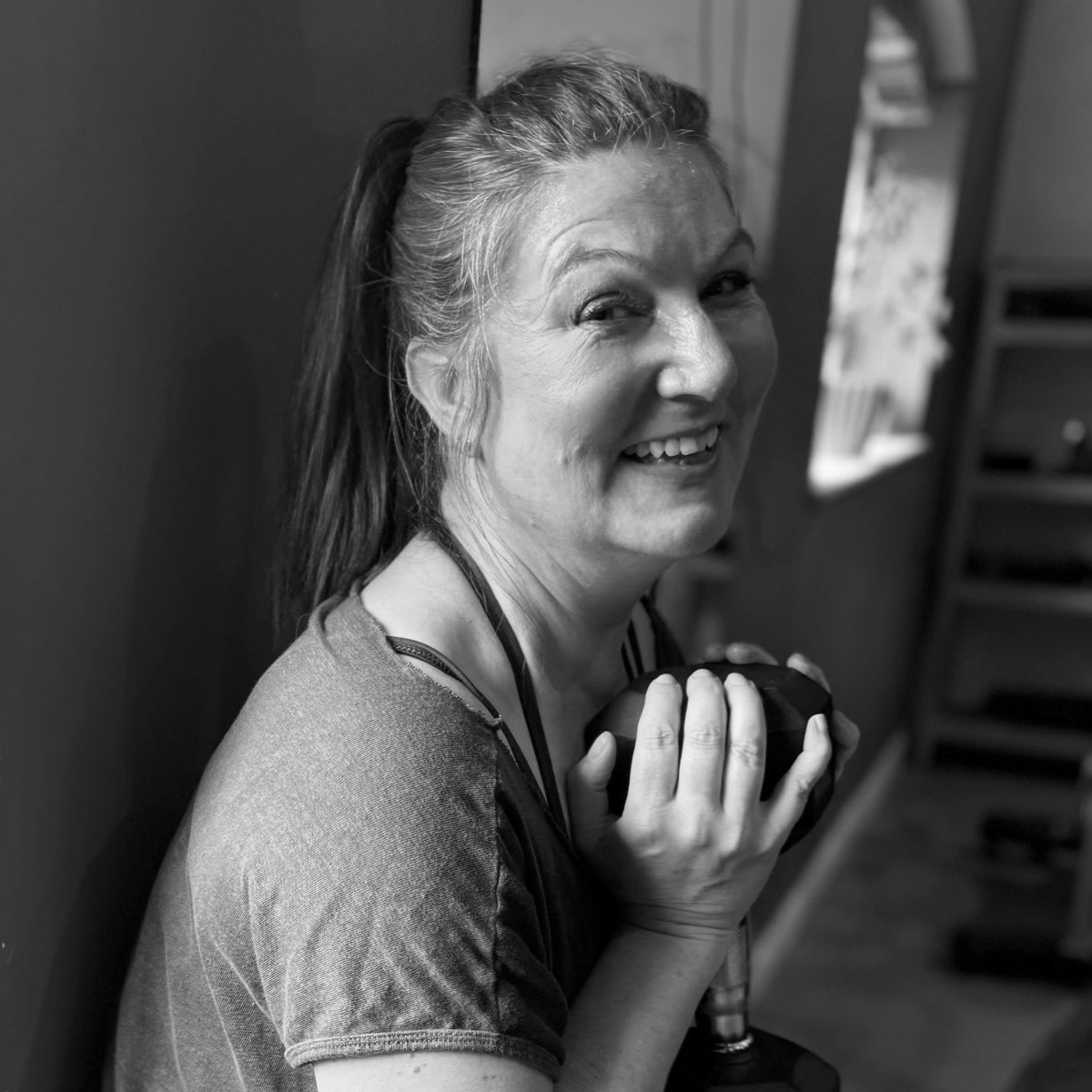 Woman smiling while holding a dumbbell, indoors, wearing a gray shirt, ponytail.