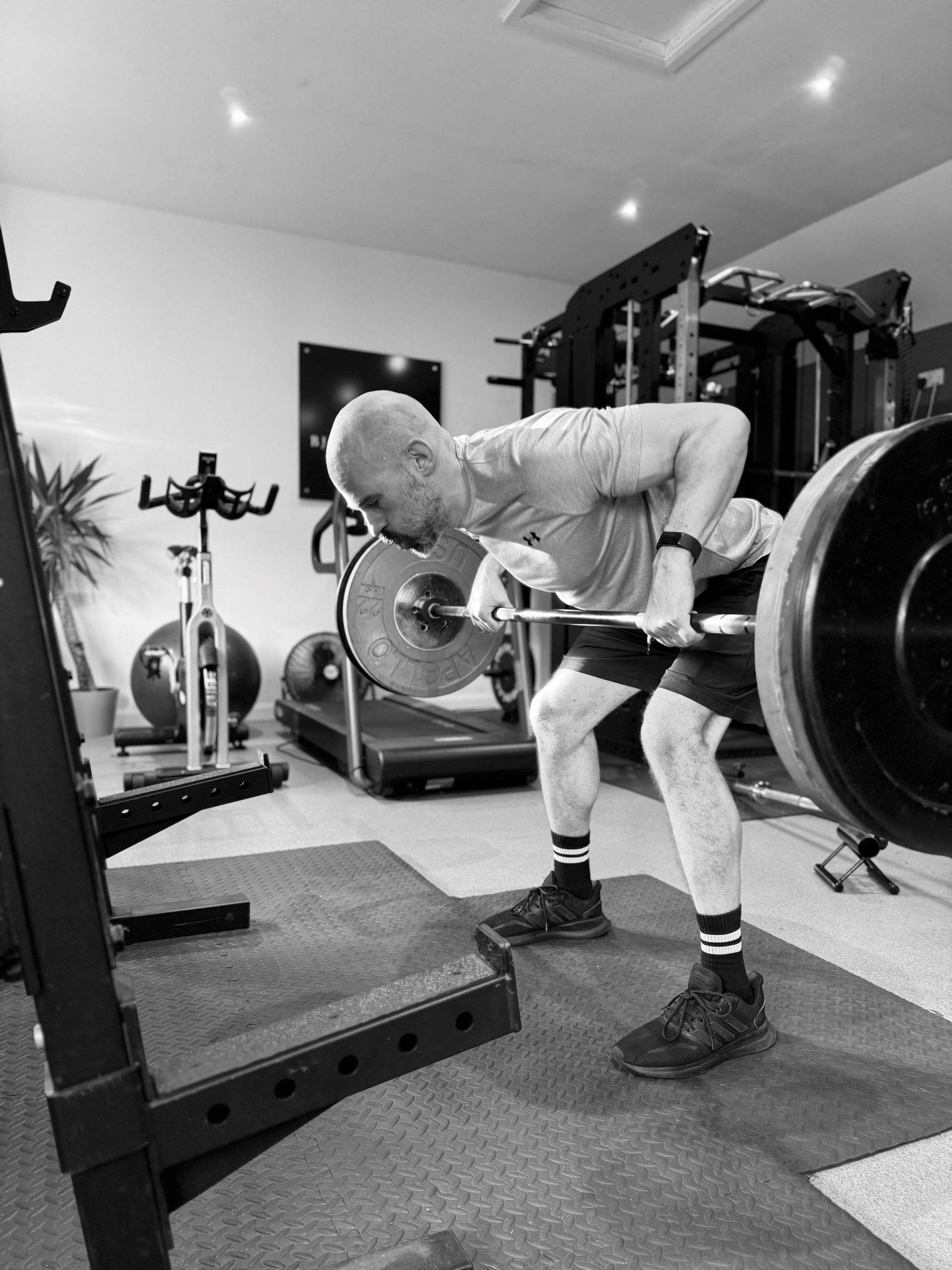 Man in a home gym, bent over, performing a barbell row exercise. Black and white.