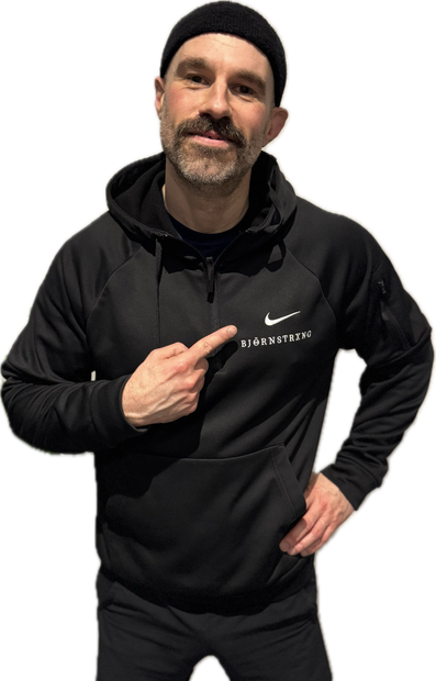 Man in black hoodie and beanie points to logo on chest.