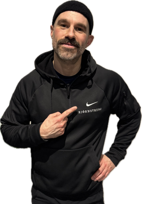 Man in black hoodie and beanie points to logo on chest.