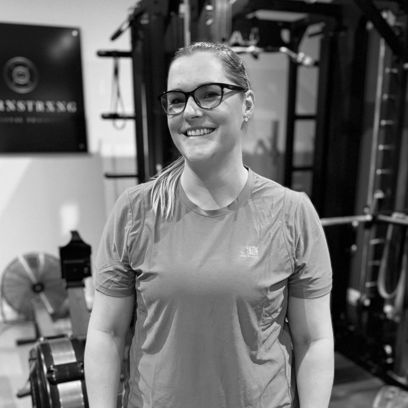 Woman with glasses smiling in a gym, wearing a t-shirt. Behind her are exercise machines.