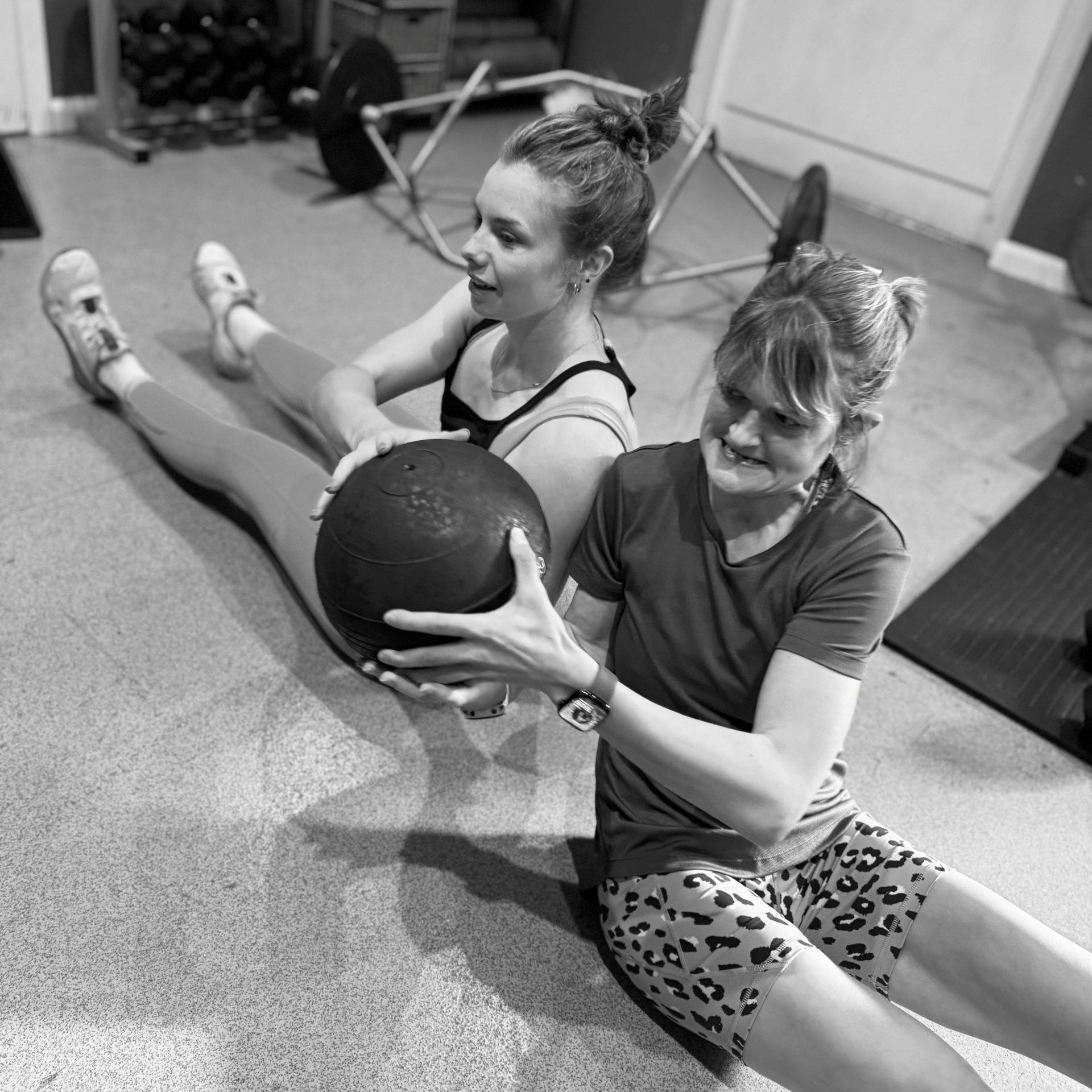 Two women exercise, seated, holding a medicine ball and twisting in a home gym.