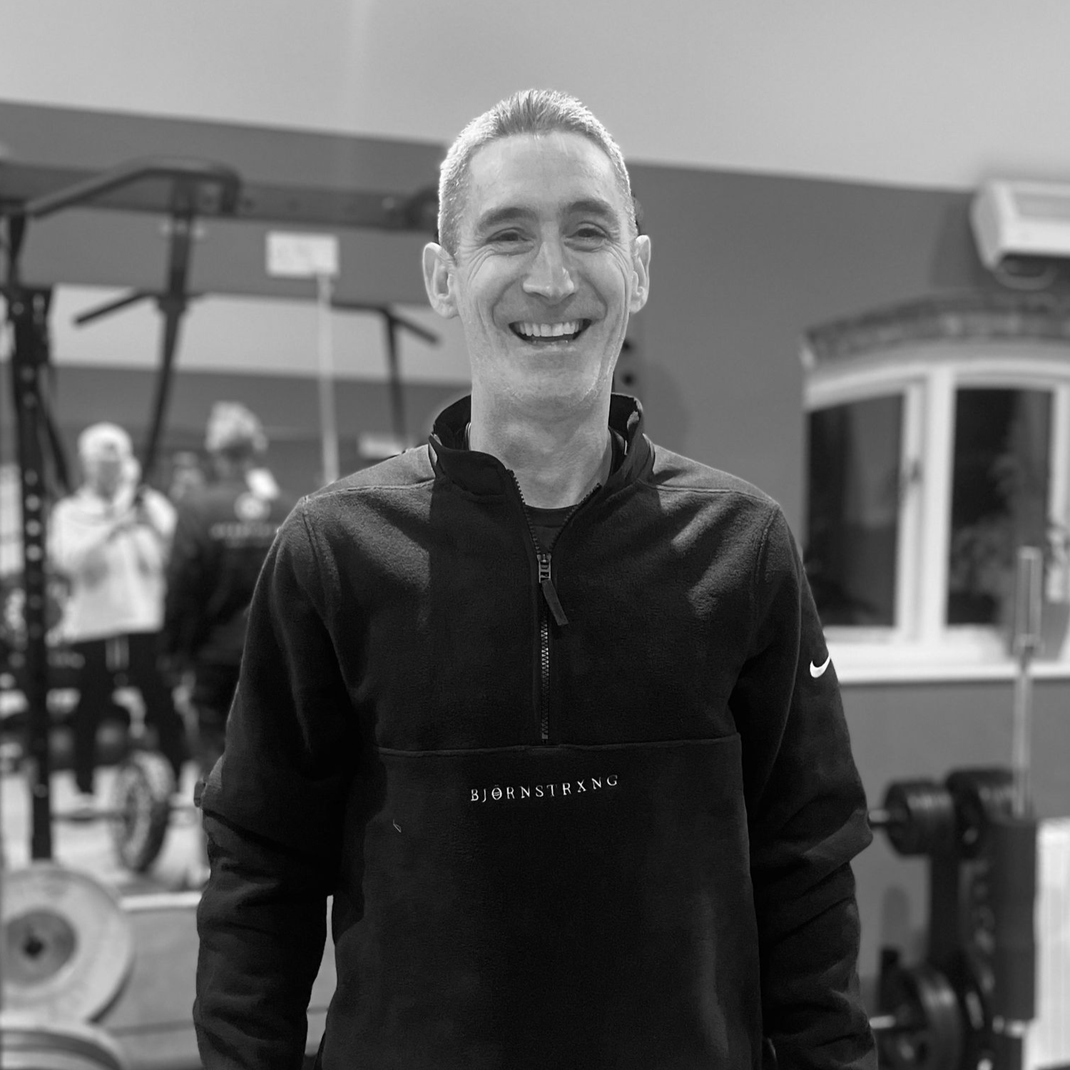 Man smiling in a gym, wearing a black sweatshirt. Weights and equipment in the background.