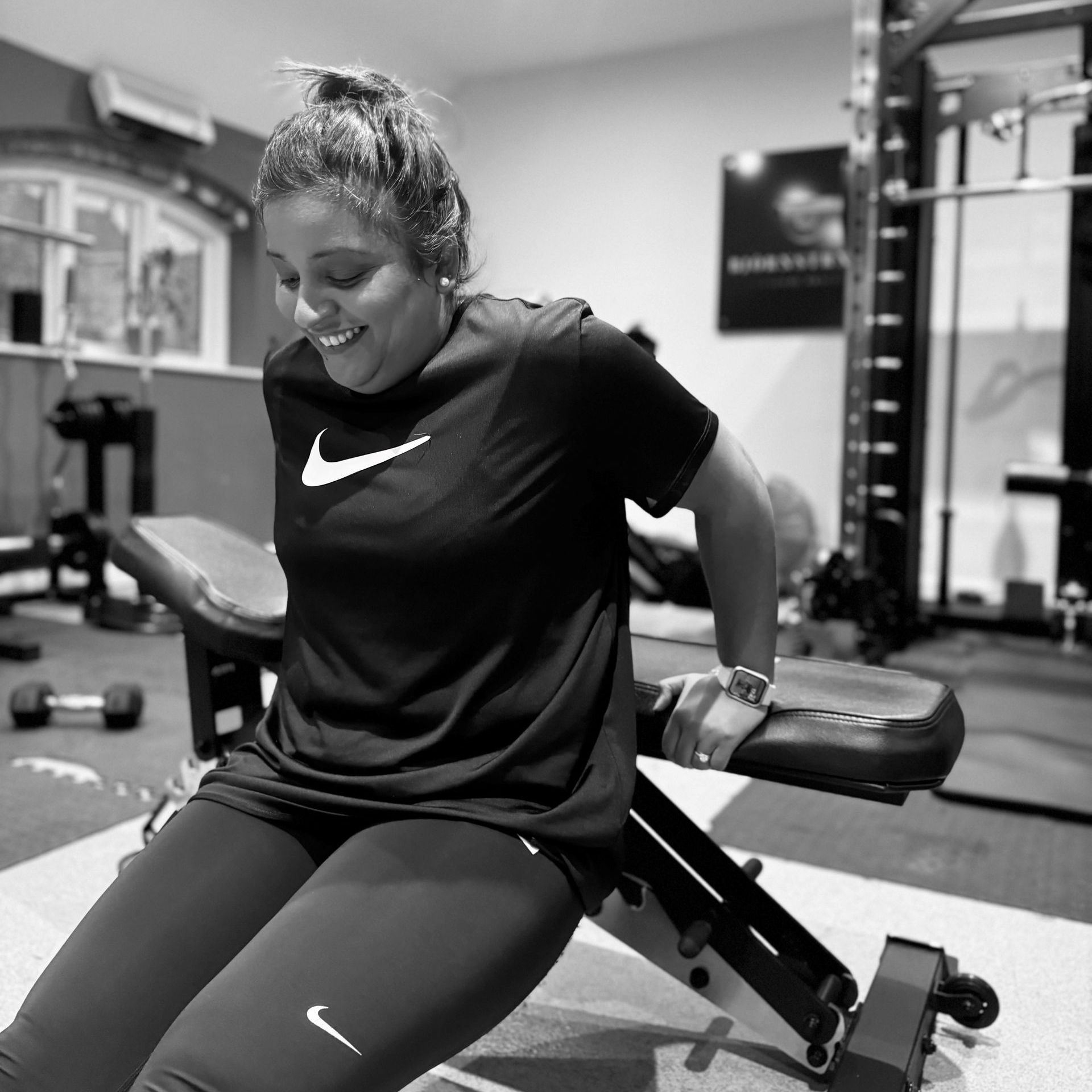 Woman doing tricep dips on a weight bench in a home gym, smiling, wearing Nike shirt and leggings.