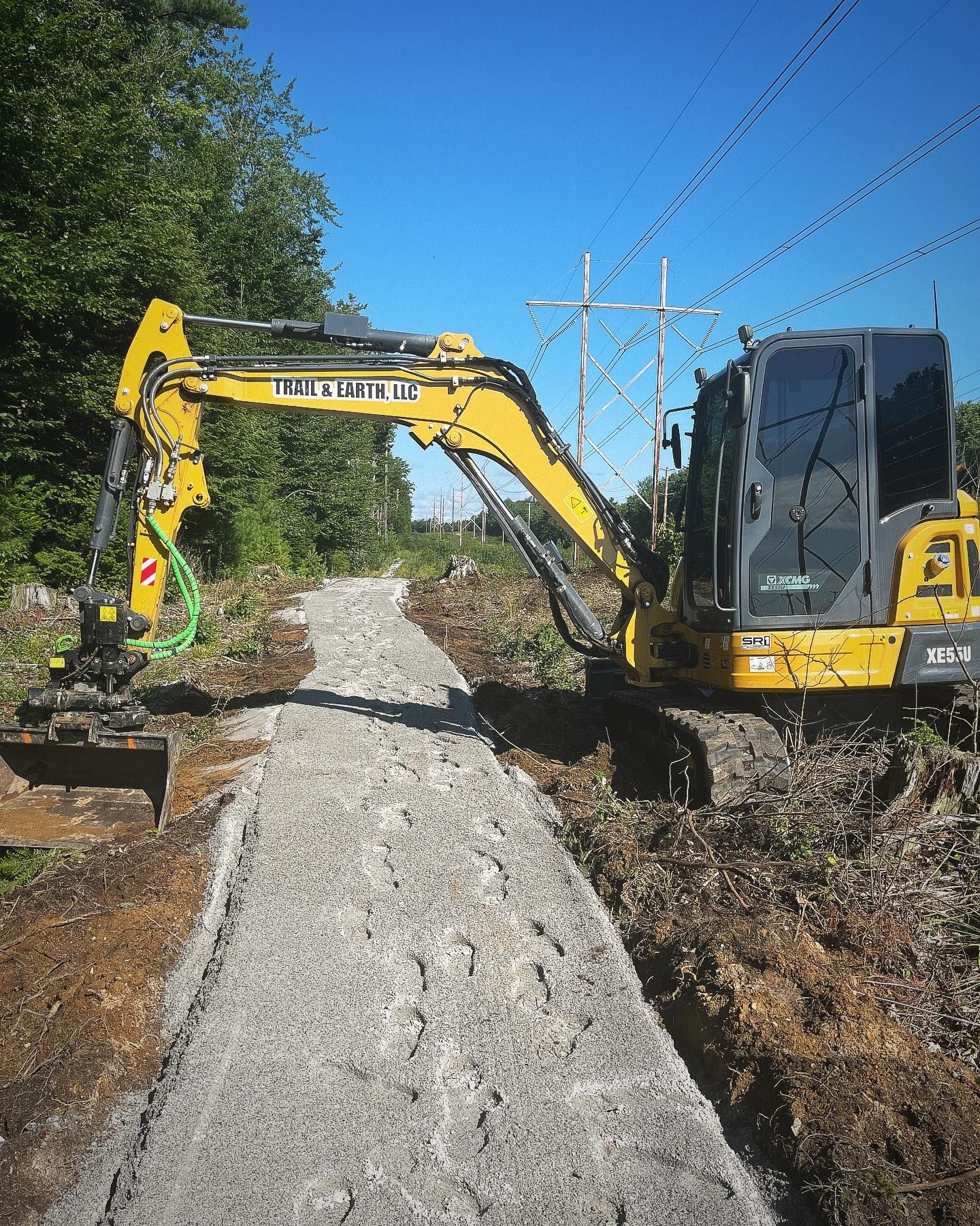 Yellow excavator constructing a gravel path alongside a treeline under a blue sky.