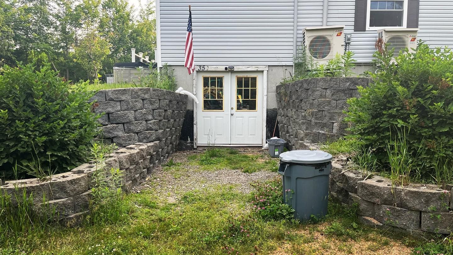 Gray stone retaining walls frame a pathway to a white door under a building. An American flag hangs above.