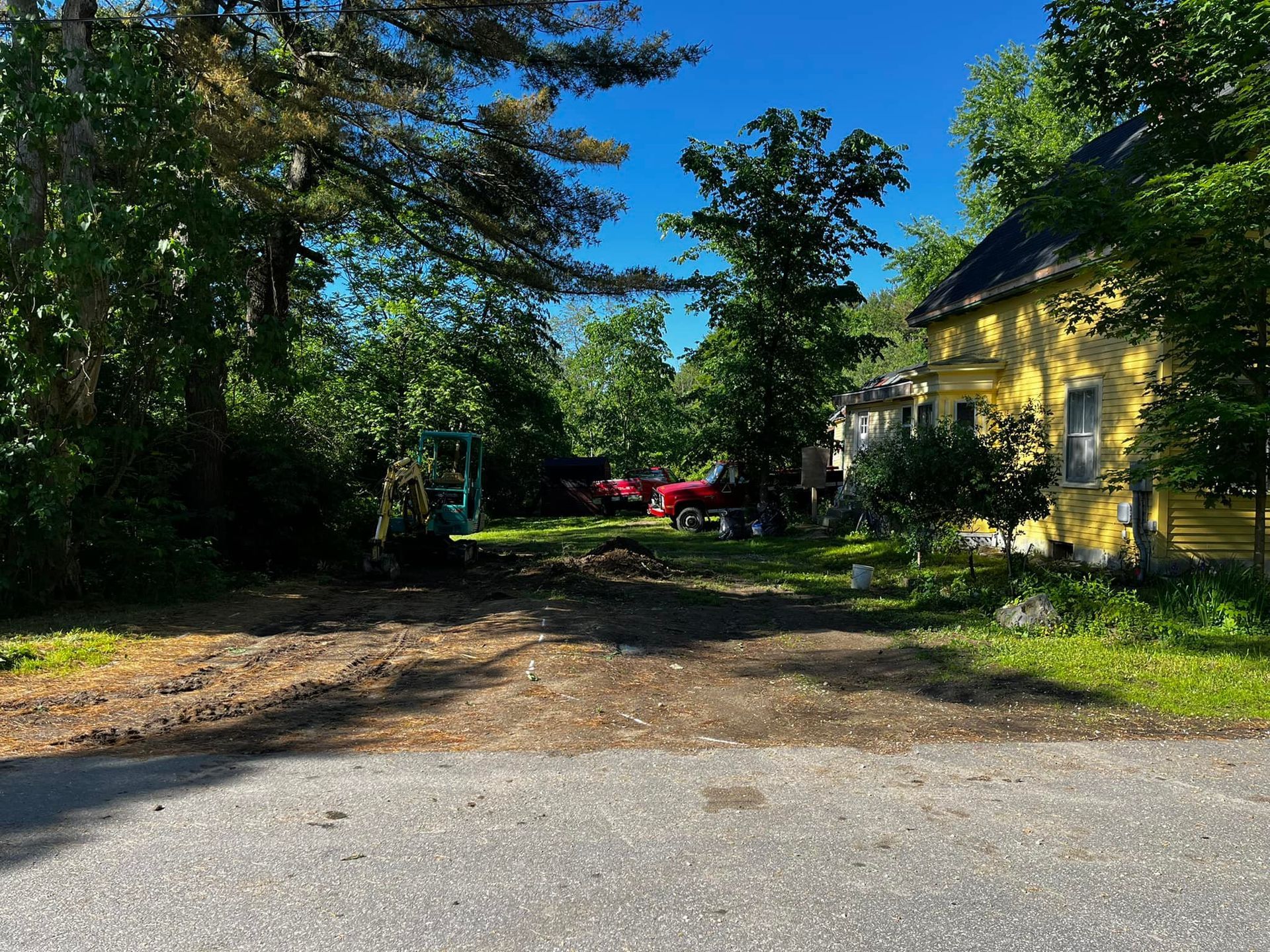 Dirt lot with trees, a yellow house, and vehicles in a sunny setting.