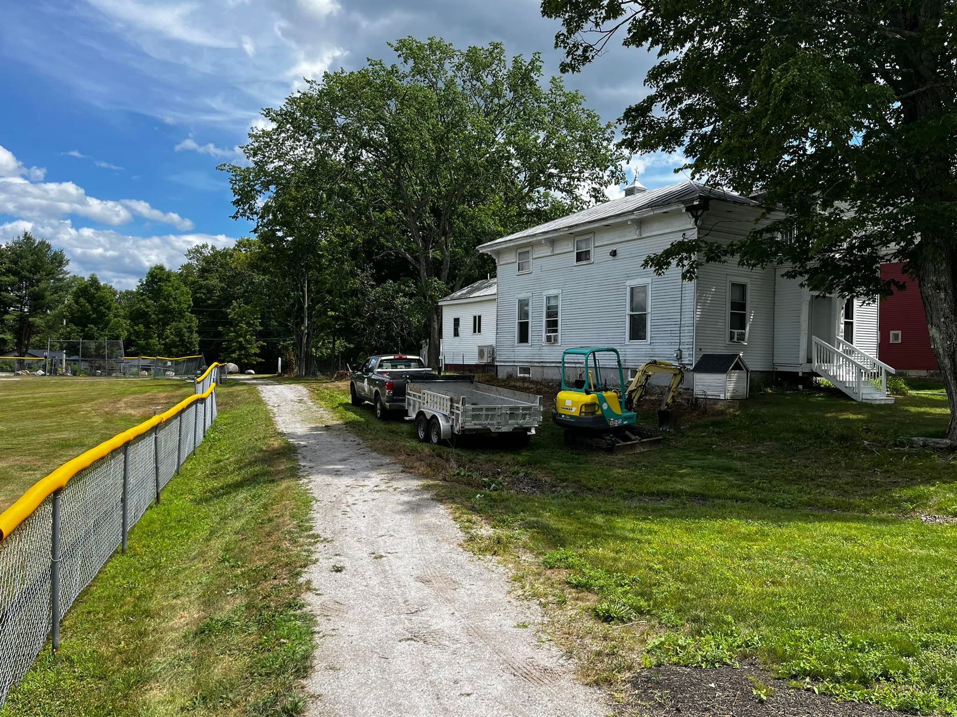 White house next to a gravel driveway, with vehicles and trees on a sunny day.