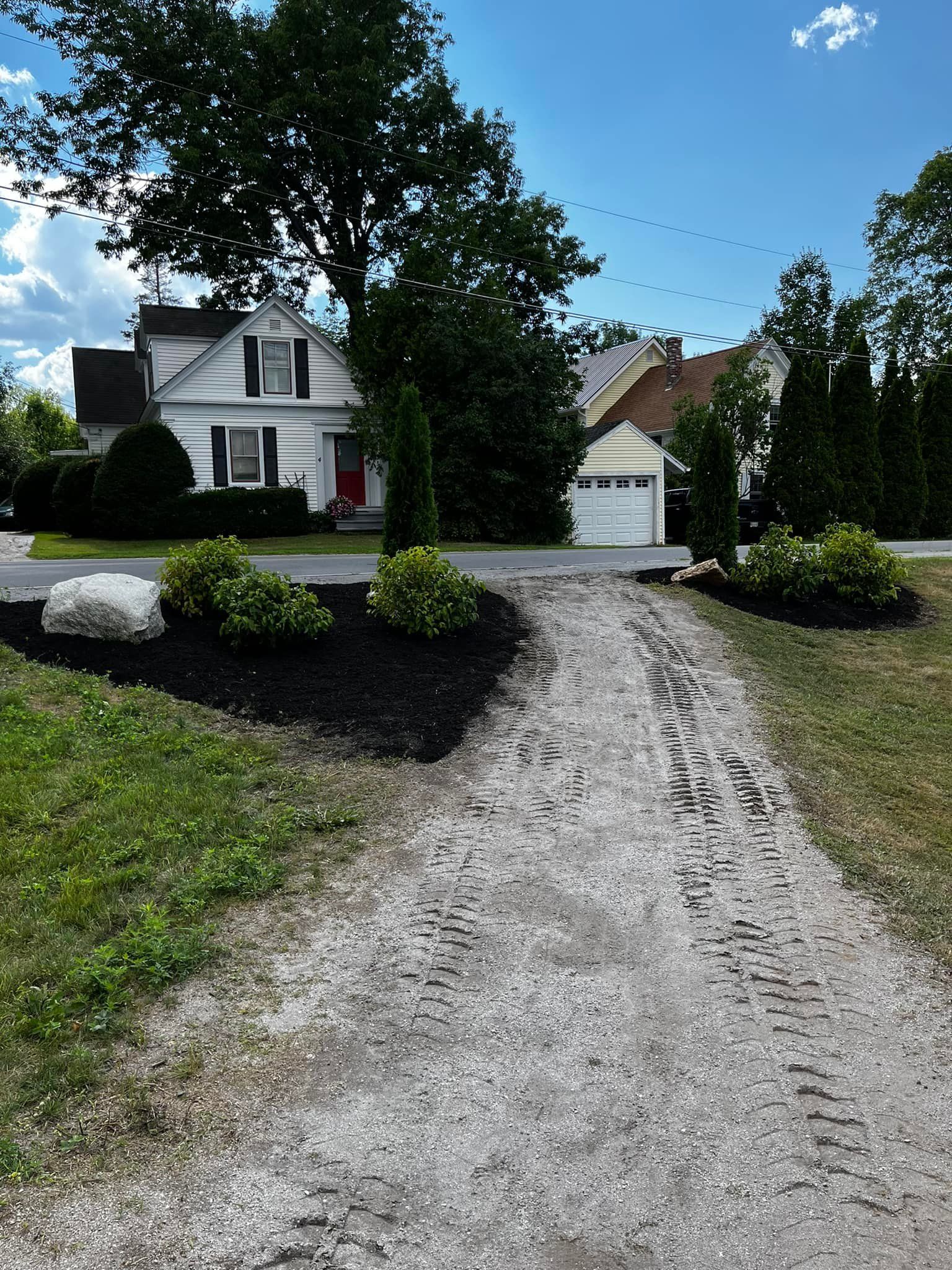 Gravel driveway leading to a white house with a black-mulched garden bed and green lawn under a blue sky.