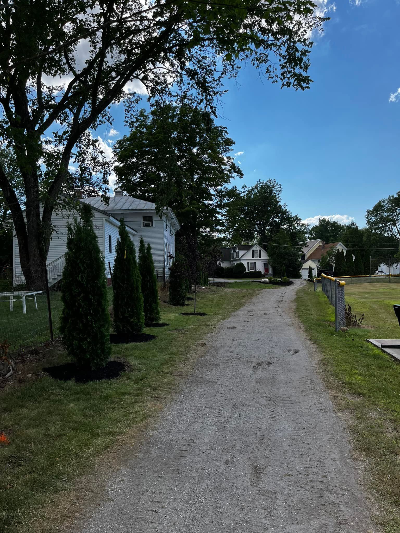 Gravel path leads to white houses under a bright blue sky with trees and evergreens.