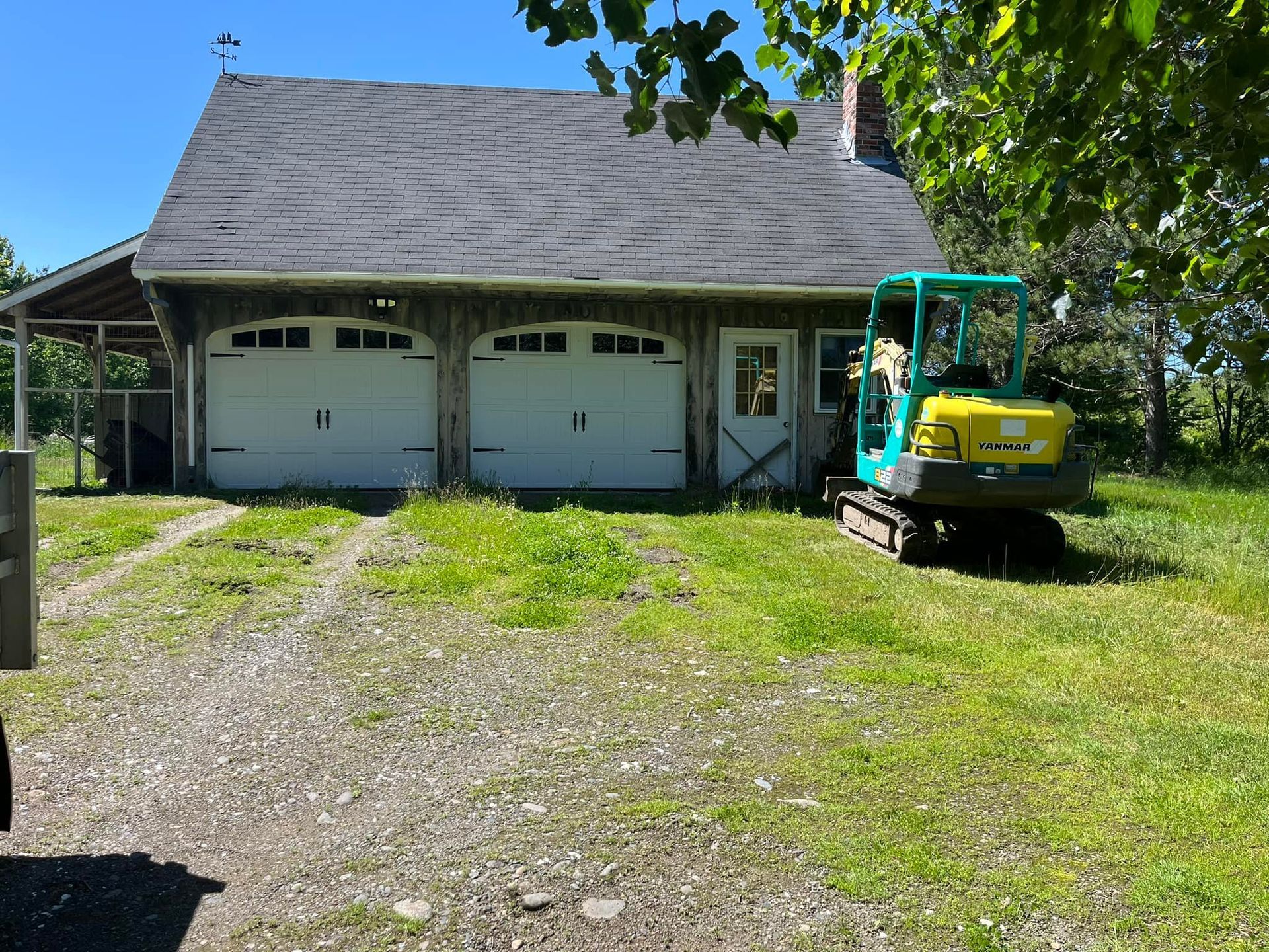 A weathered building with two garage doors and a small excavator parked outside on a grassy lawn.