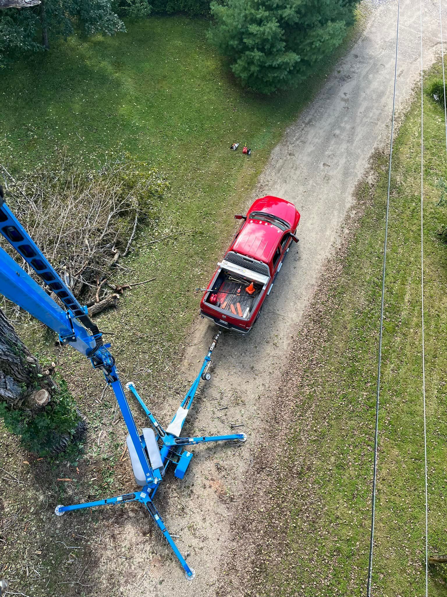 Red pickup truck being towed by a blue crane on a dirt road, viewed from above. Green grass and trees surround.