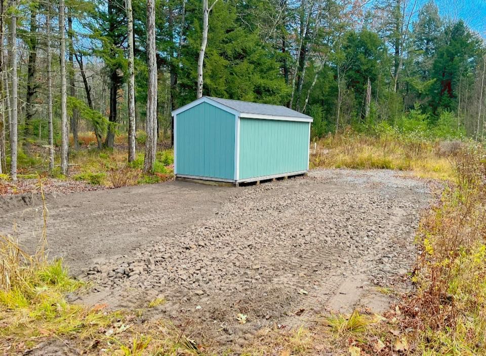 Blue shed on a gravel base in a wooded area.