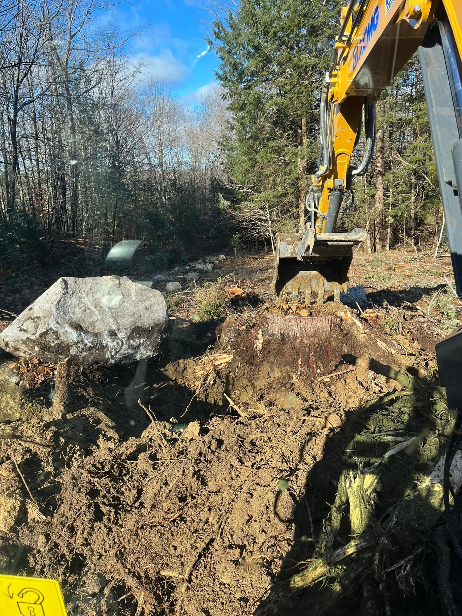 An excavator digs into dirt in a wooded area, near a large boulder and trees under a blue sky.