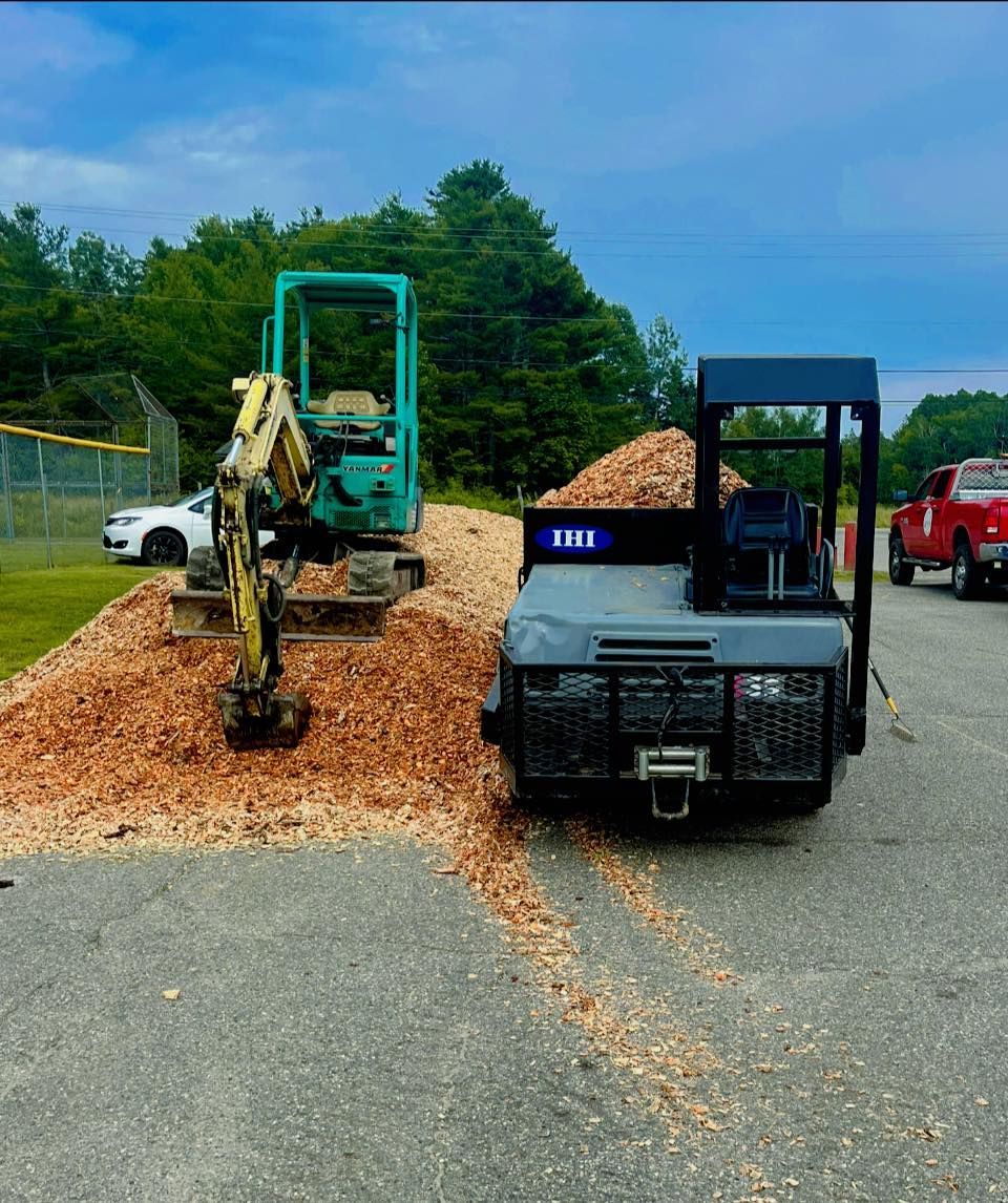 Mini excavator and small hauler on a pile of wood chips, at a ball field.