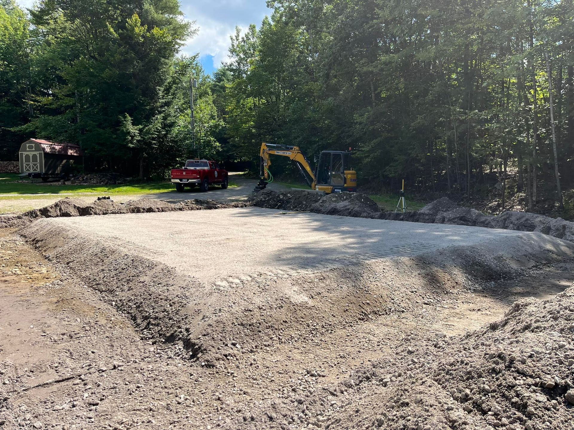 Construction site with a gravel base, an excavator, and a red pickup truck. Trees in background.
