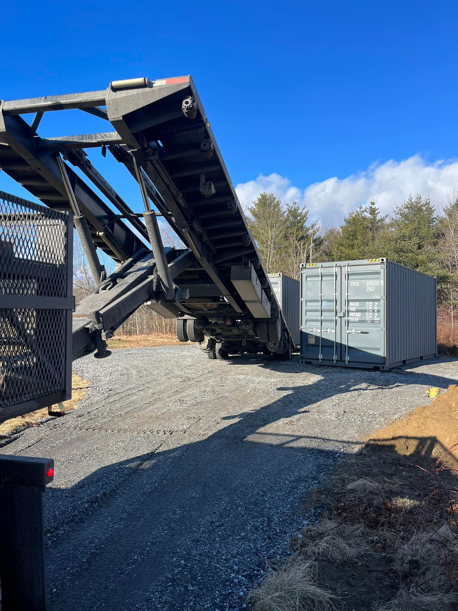 A cargo container being unloaded from a truck with a ramp on a gravel driveway under a blue sky.
