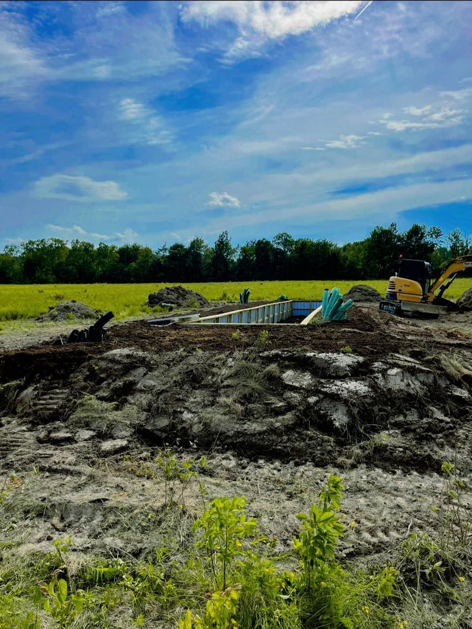 Construction site with an excavator, circular foundation, and piles of dirt under a blue sky.