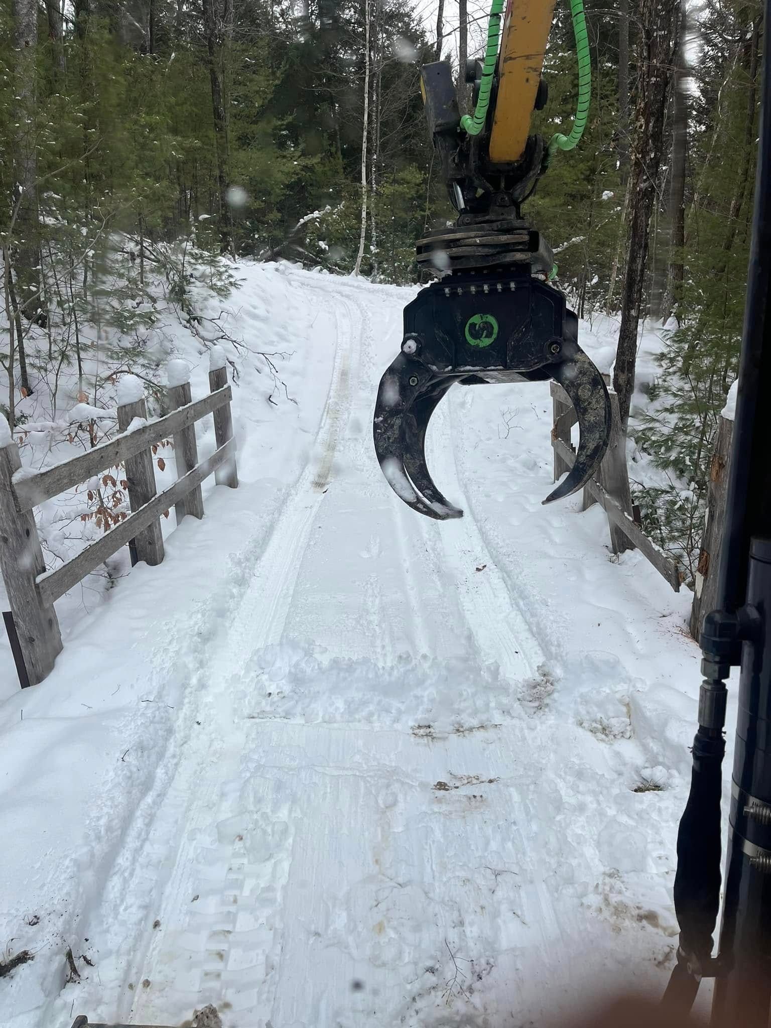 Snowy road with an excavator claw, flanked by a wooden fence, leading into a forest.