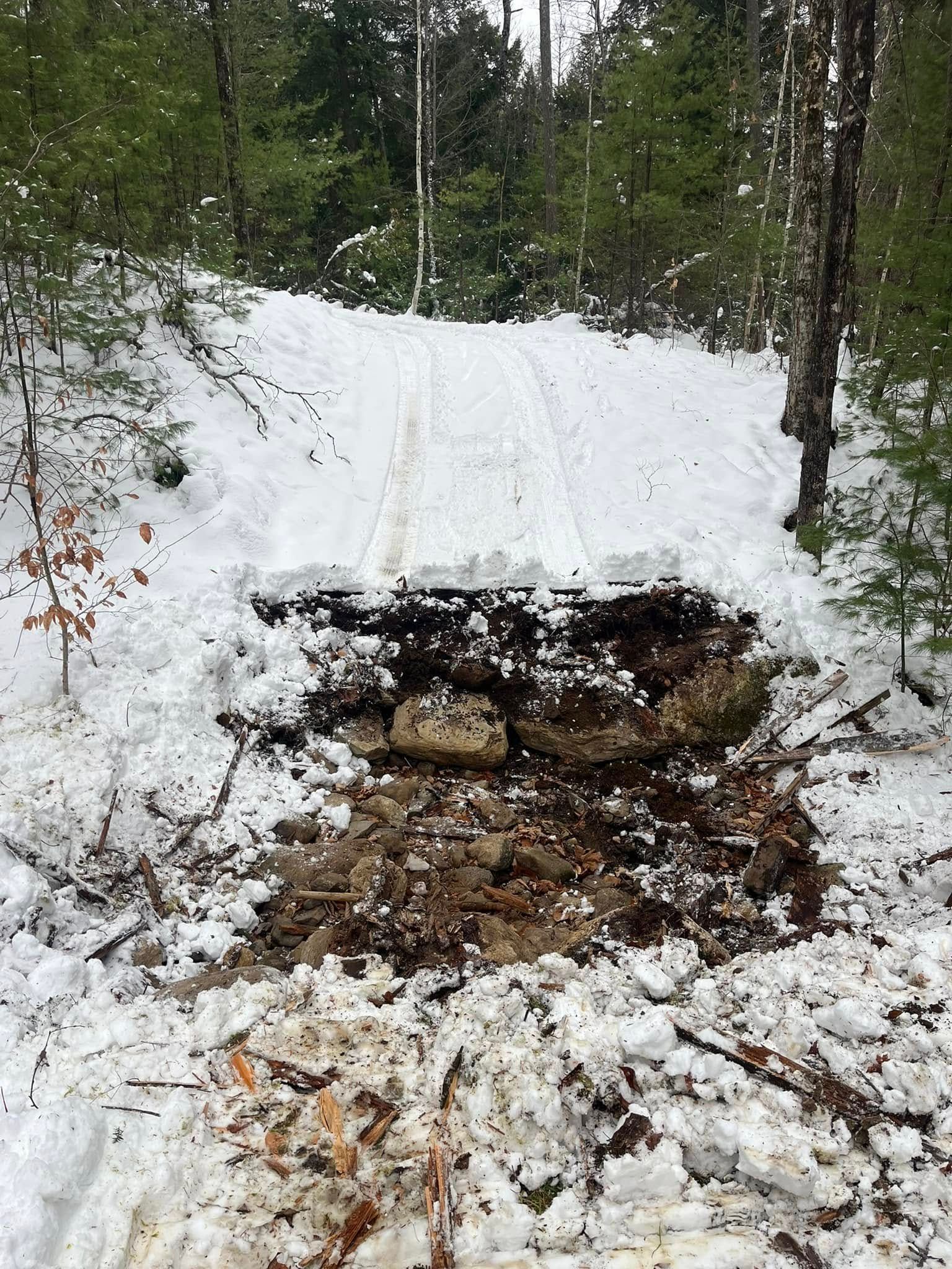 Snow-covered path in a forest, partially blocked by a snow and dirt obstruction.