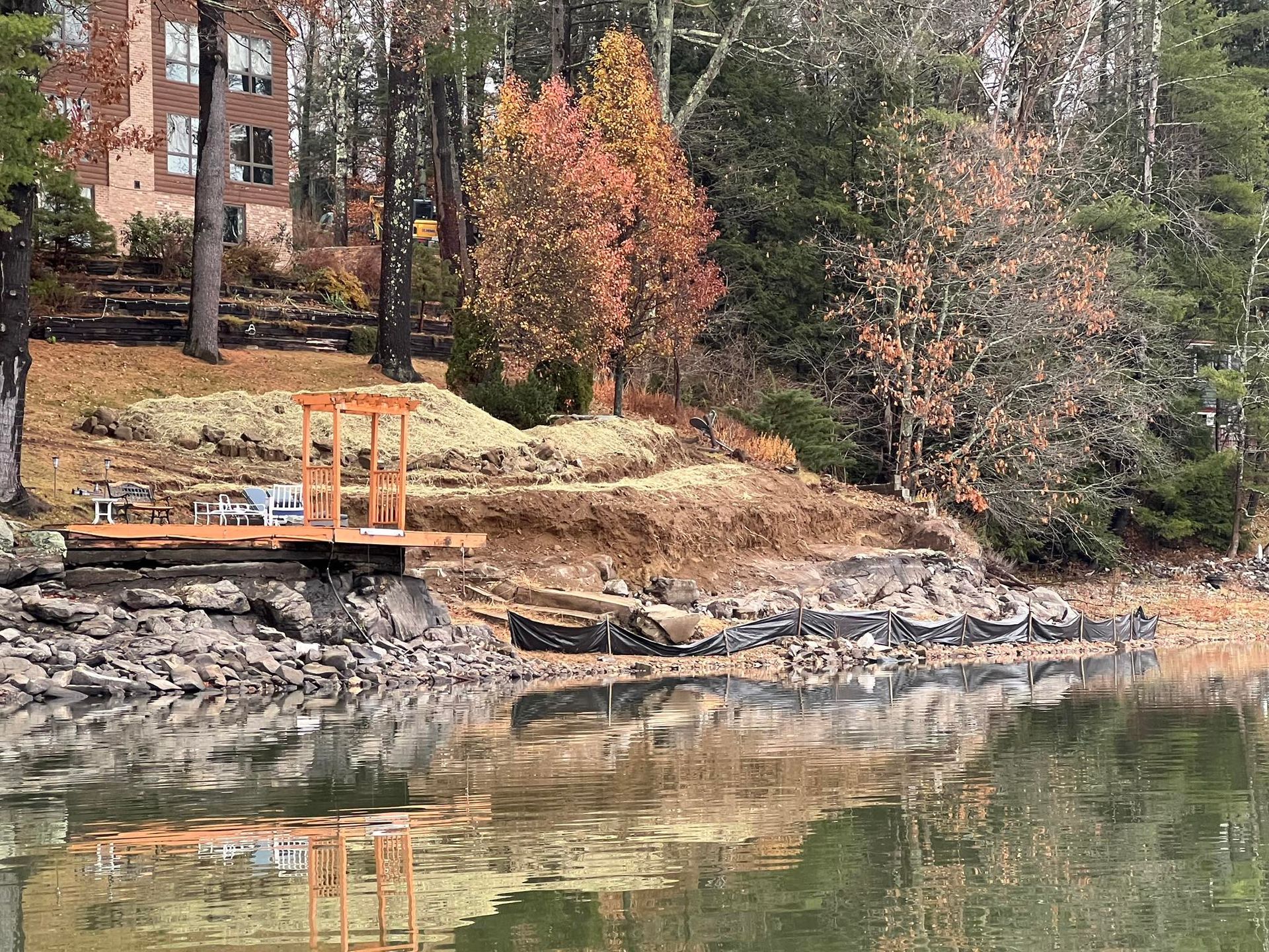 Dock with pergola over rocky shoreline, autumn foliage, lake water. House in background.
