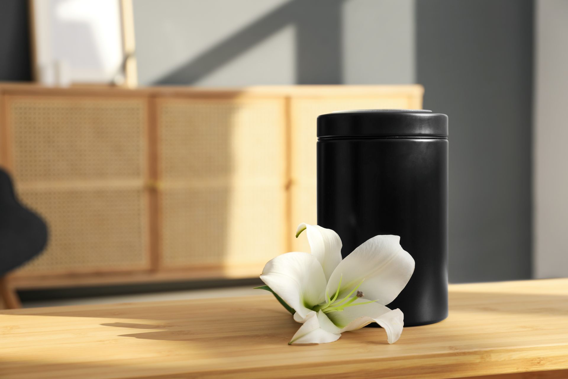 Black urn and white flower on wooden table, cabinet in background.