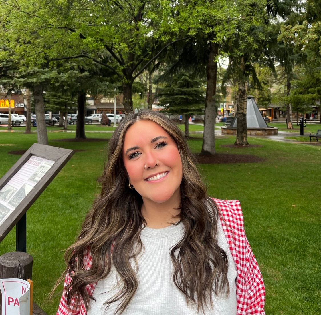 Woman with long dark hair, glasses, and floral top smiles in front of a brick wall and a large metal letter