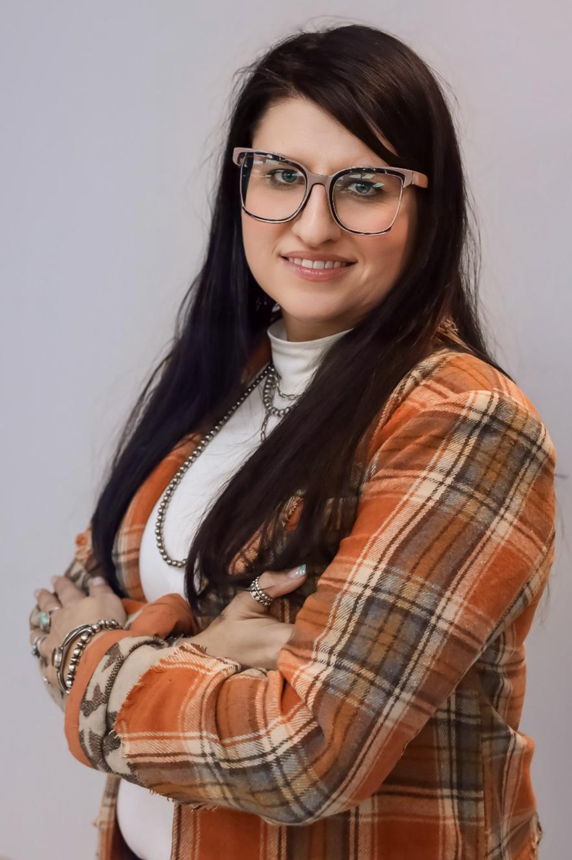 Woman with glasses and arms crossed, wearing a plaid jacket and white turtleneck, smiling.